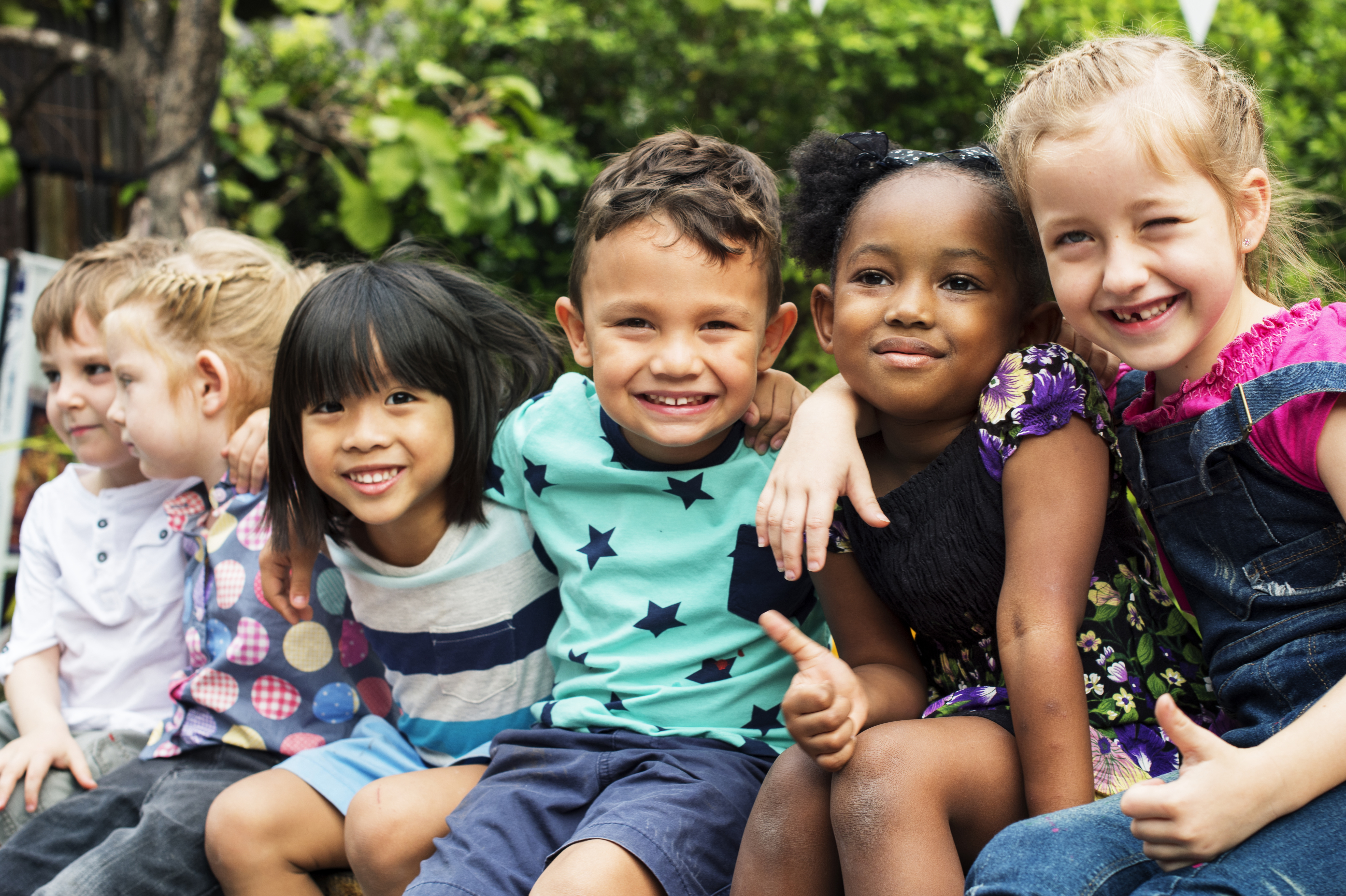 6 kids sitting outdoors with trees in the background. The kids have arms around each other and are smiling at the camera.