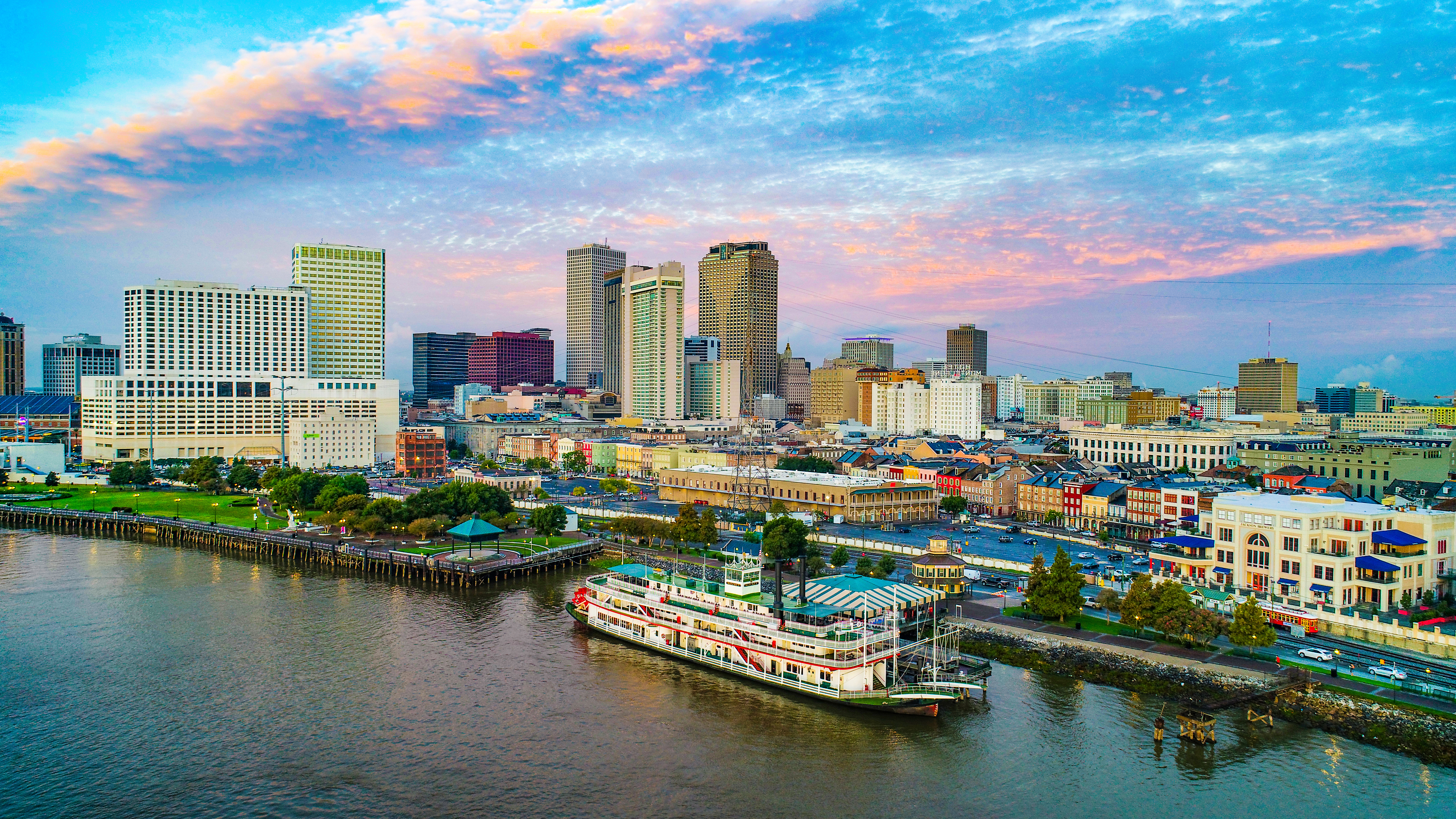A photograph of New Orleans. The top third is the sky with a light punk sunset across clouds. The middle third is the New Orleans skyline with several skyscrapers and smaller buildings. The lowest third is a river with a large boat in it.