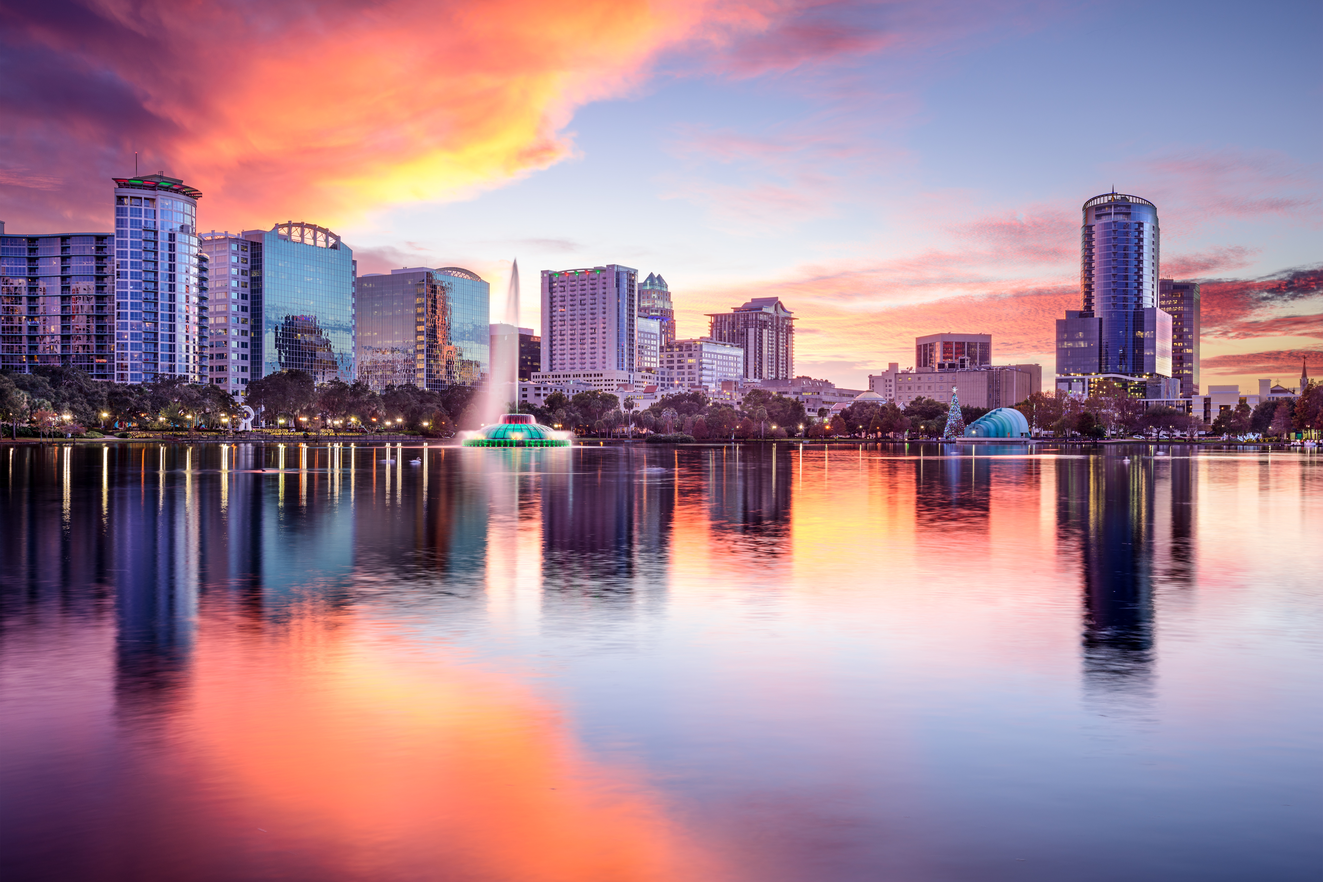A skyline of Orlando, Florida at sunset. There is a row of buildings with a purple and orange sunset above. The sunset reflects onto a lake, which is front of the buildings in the foreground.