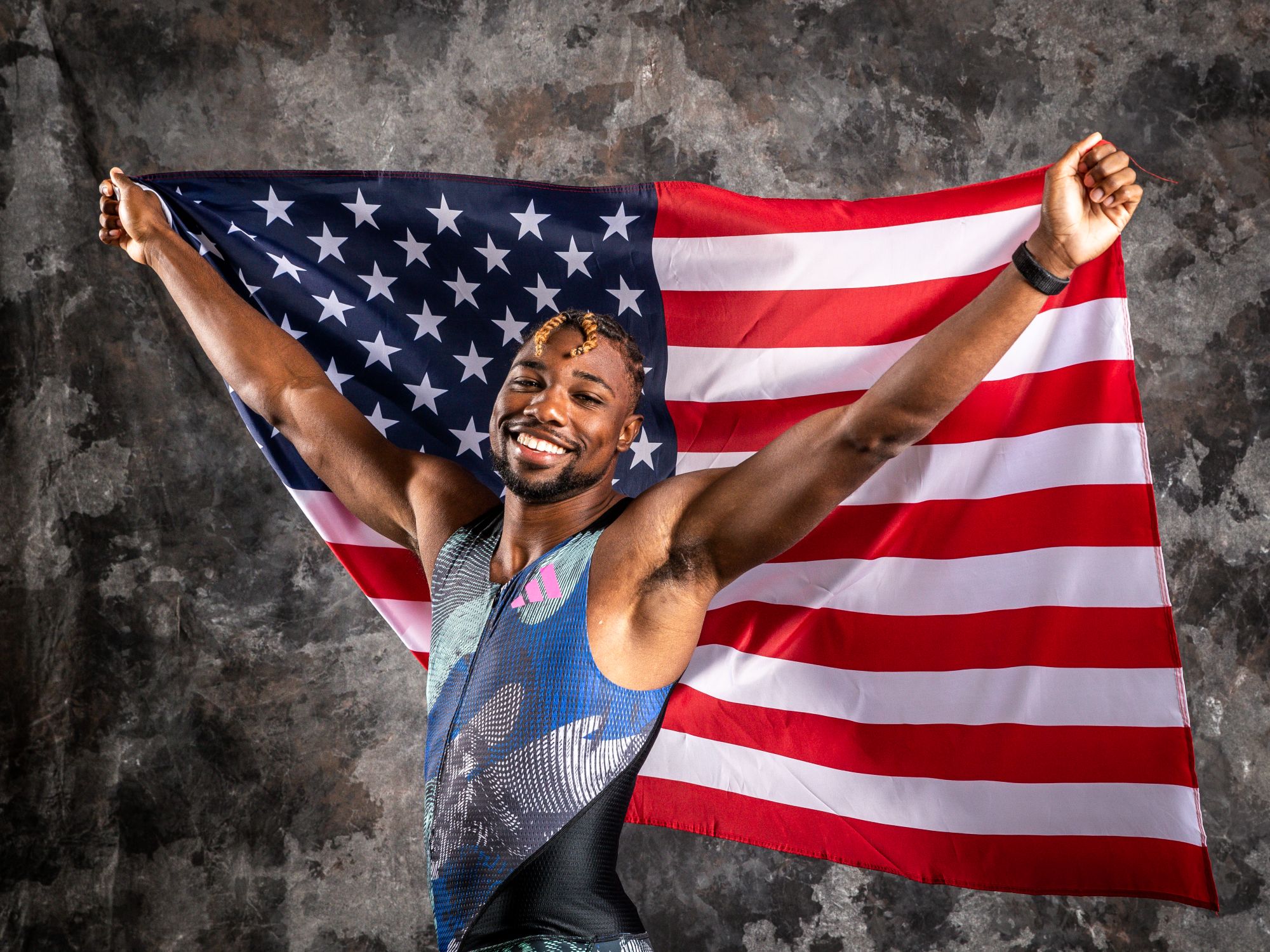 Olympic athlete Noah Lyles stands smiling and holding a USA flag above his head.